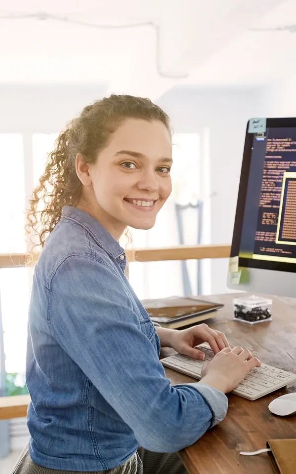 Porträt einer IT-Entwicklerin, die in die Kamera lächelt, während sie auf der Tastatur tippt, mit Programmiercode auf dem Computerbildschirm